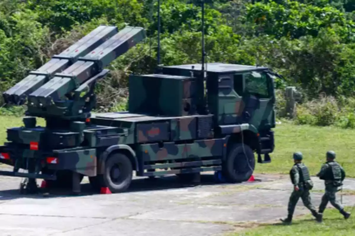 Camouflaged military vehicle equipped with missile launchers and two soldiers walking nearby in an outdoor environment with vegetation in the background