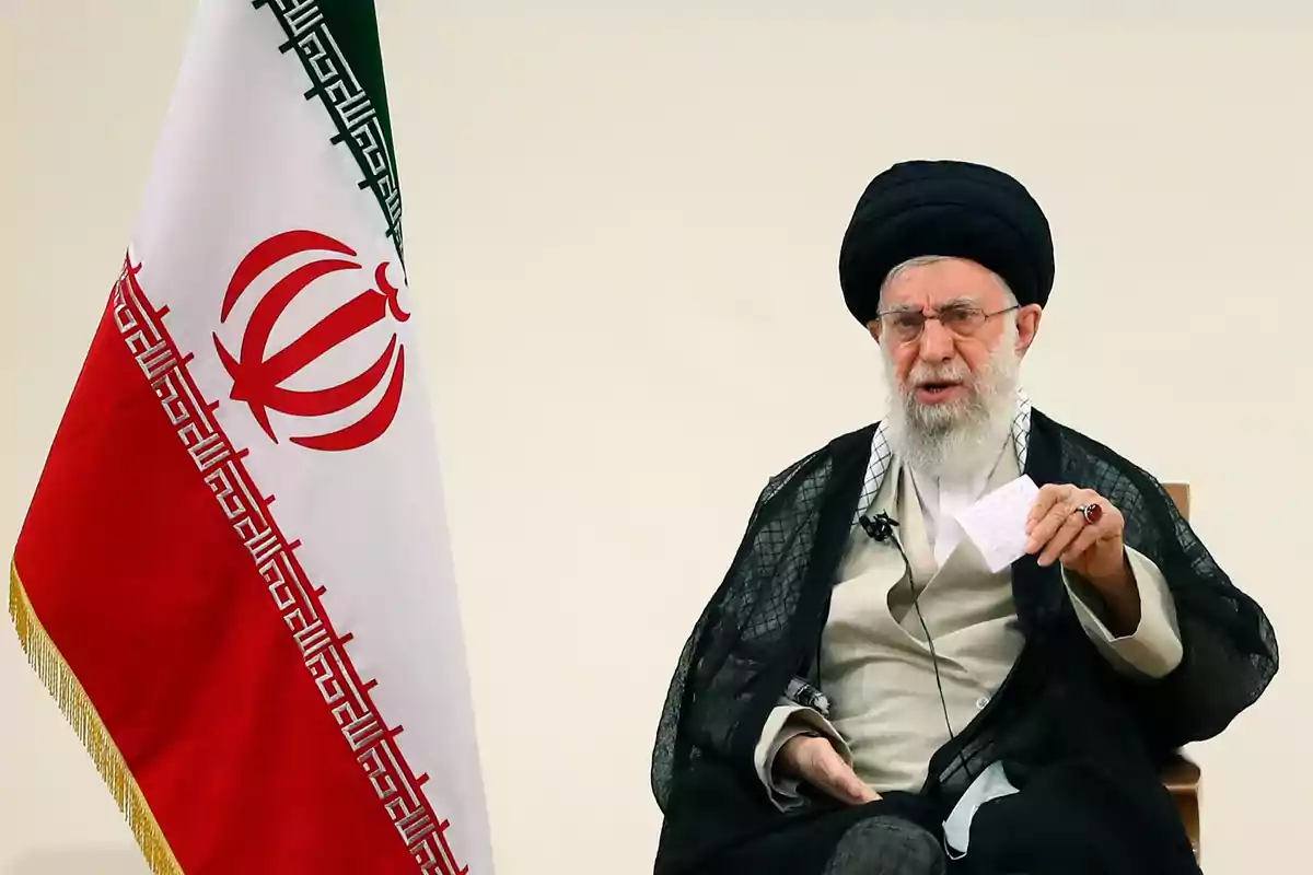 Hombre mayor con barba y turbante negro sentado junto a una bandera de Irán sosteniendo una hoja de papel Hombre mayor con barba y turbante negro sentado junto a una bandera de Irán sosteniendo una hoja de papel