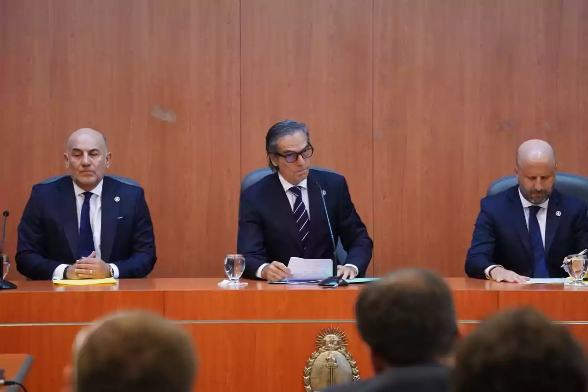 Three men in dark suits sitting at a wooden table during a formal meeting, with microphones and glasses of water in front of them and a national emblem in the center of the table. Three men in dark suits sitting at a wooden table during a formal meeting, with microphones and glasses of water in front of them and a national emblem in the center of the table.