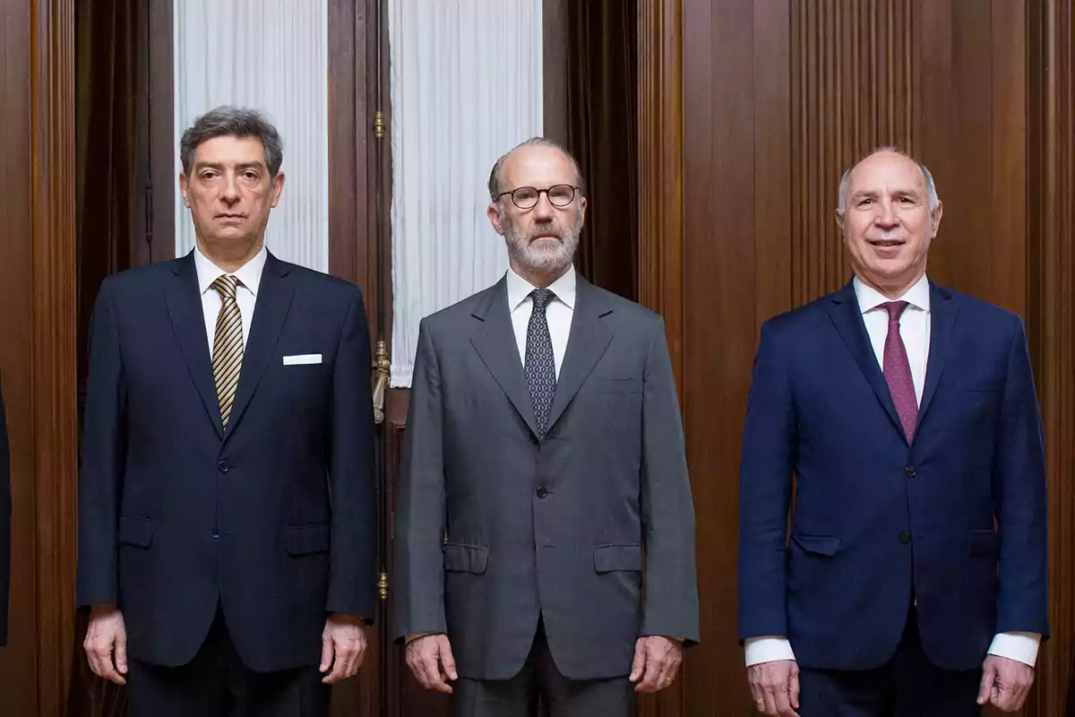Tres hombres de traje posan de pie en una sala con paredes de madera