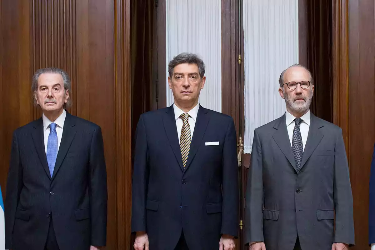 Tres hombres de pie en trajes formales frente a una puerta de madera con cortinas blancas.