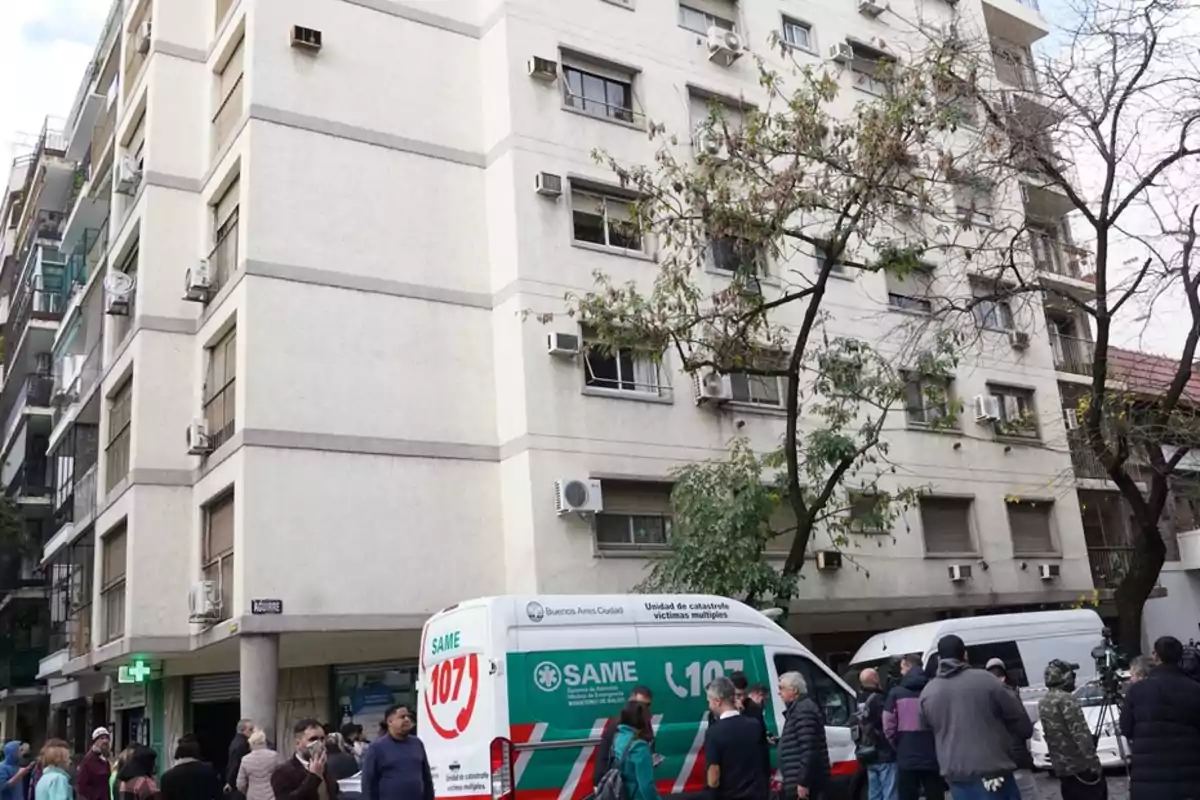 Un edificio de apartamentos con varias unidades de aire acondicionado en la fachada y un árbol en primer plano; en la calle hay una ambulancia del SAME y varias personas reunidas. Un edificio de apartamentos con varias unidades de aire acondicionado en la fachada y un árbol en primer plano; en la calle hay una ambulancia del SAME y varias personas reunidas.