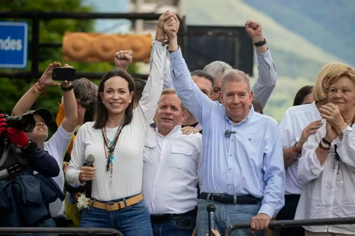 Un grupo de personas vestidas de blanco y azul se encuentra en un evento al aire libre, dos de ellas levantan las manos unidas en señal de celebración mientras otras personas a su alrededor aplauden y toman fotos.