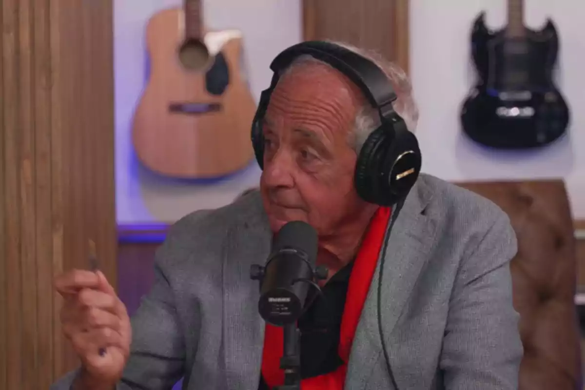 An older man with headphones and a microphone in front of him, sitting in a studio with guitars hanging on the wall. An older man with headphones and a microphone in front of him, sitting in a studio with guitars hanging on the wall.