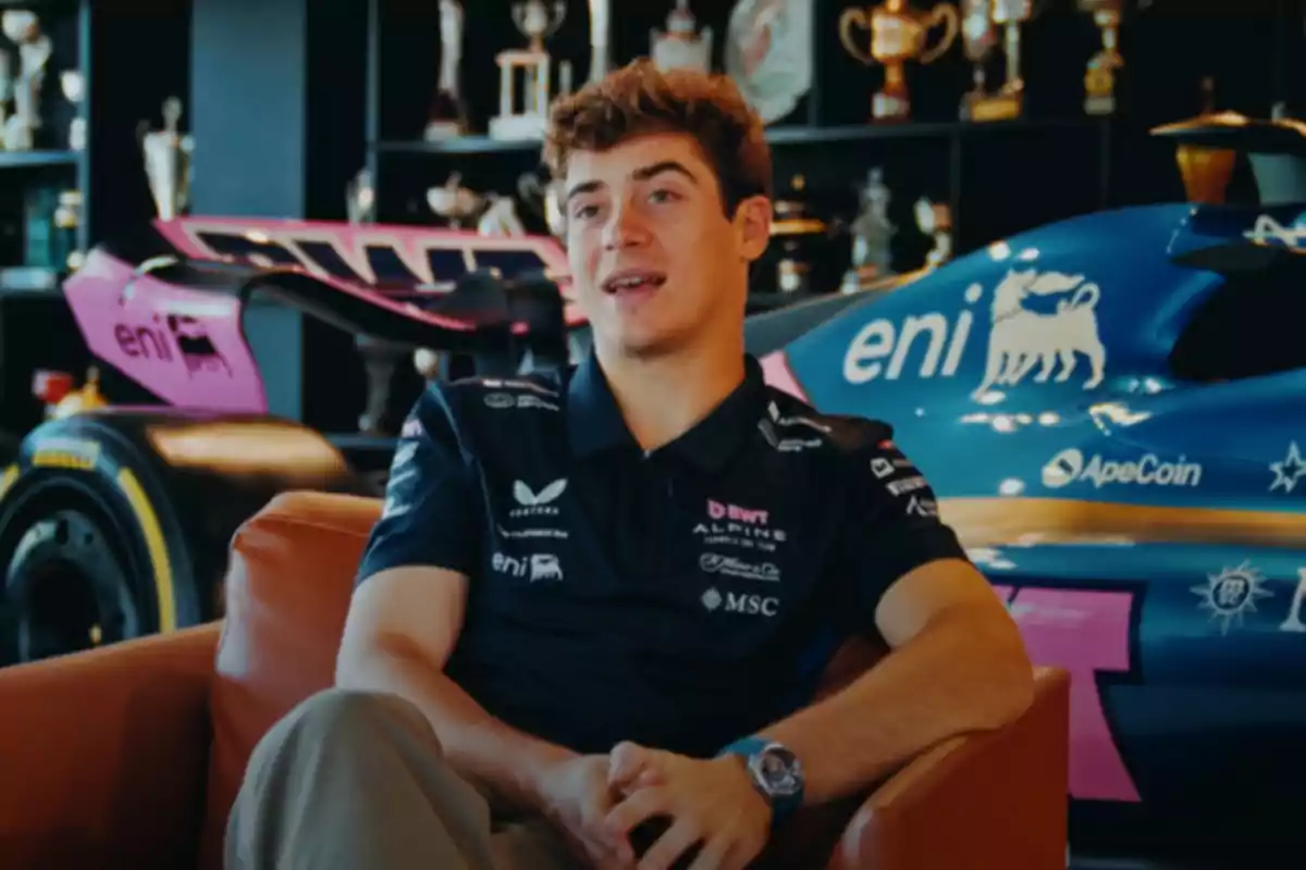 Un hombre joven sentado en un sillón, vistiendo una camiseta con logotipos de patrocinadores, con un coche de carreras y trofeos en el fondo.
