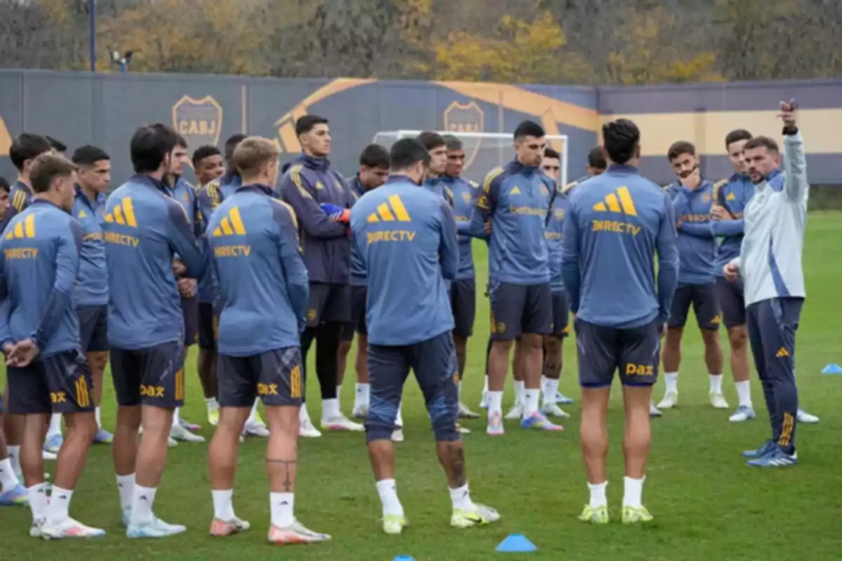 A group of players in blue and yellow training gear listens to instructions from a coach on a grass field.