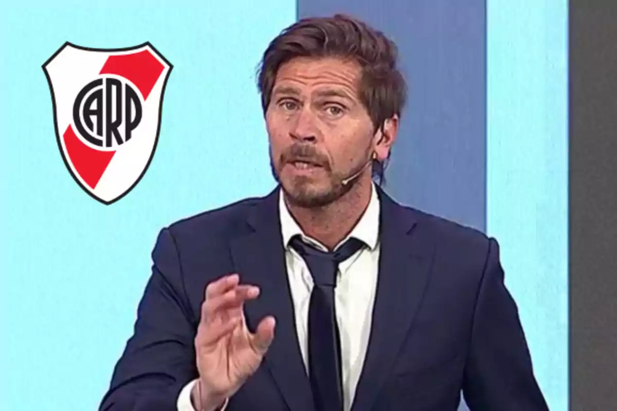 Un hombre con traje y corbata habla mientras lleva un micrófono en el oído, junto al escudo de un club de fútbol.