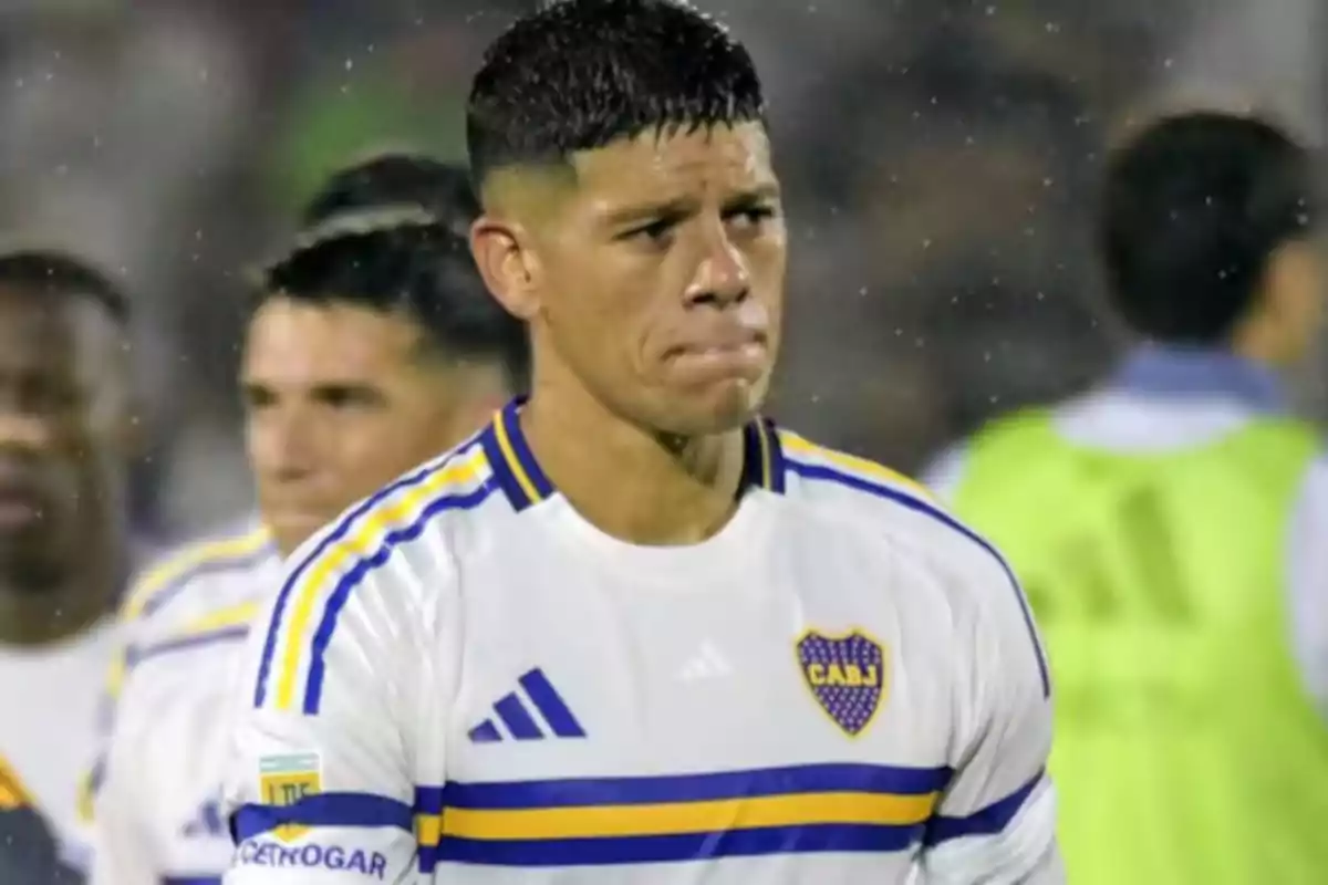 Un jugador de fútbol con expresión seria lleva una camiseta blanca con detalles azules y amarillos. Un jugador de fútbol con expresión seria lleva una camiseta blanca con detalles azules y amarillos.