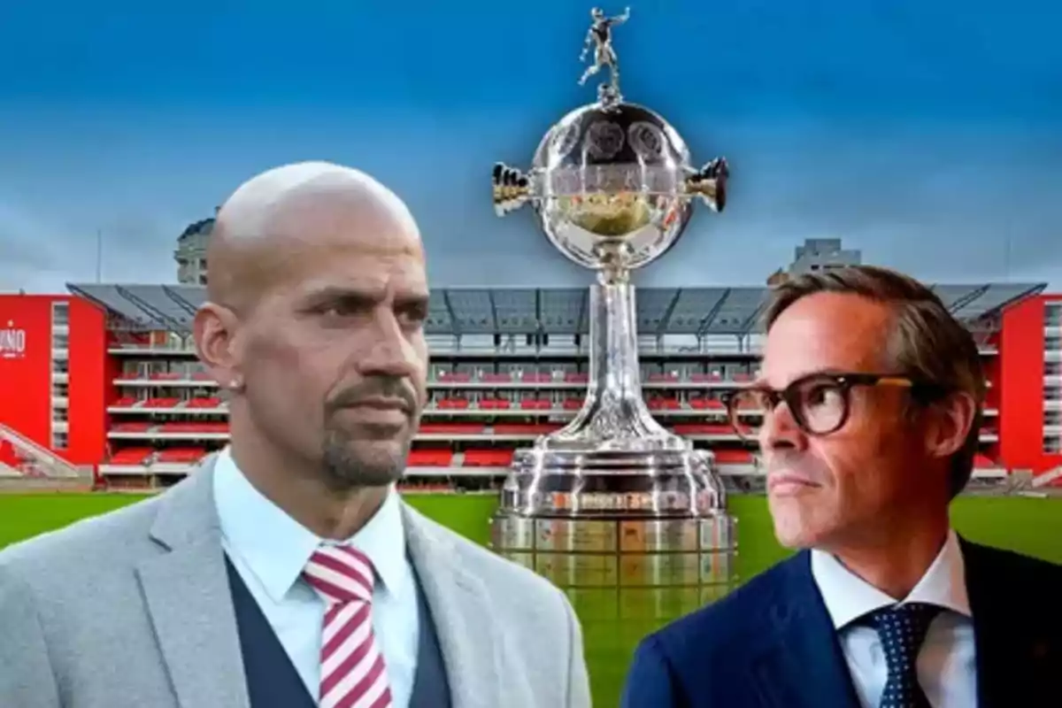 Dos hombres con trajes formales frente a un estadio de fútbol y un trofeo grande en el centro.