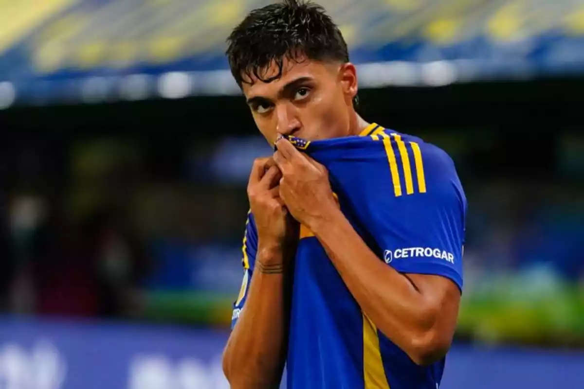 Un jugador de fútbol besa el escudo de su camiseta azul con detalles amarillos en un estadio.