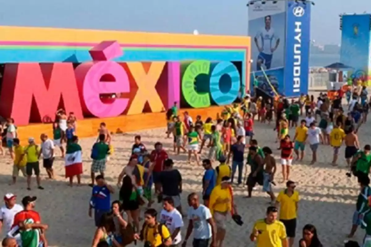 A group of people walks along a beach in front of a colorful sign that says "Mexico" with a Hyundai billboard in the background.