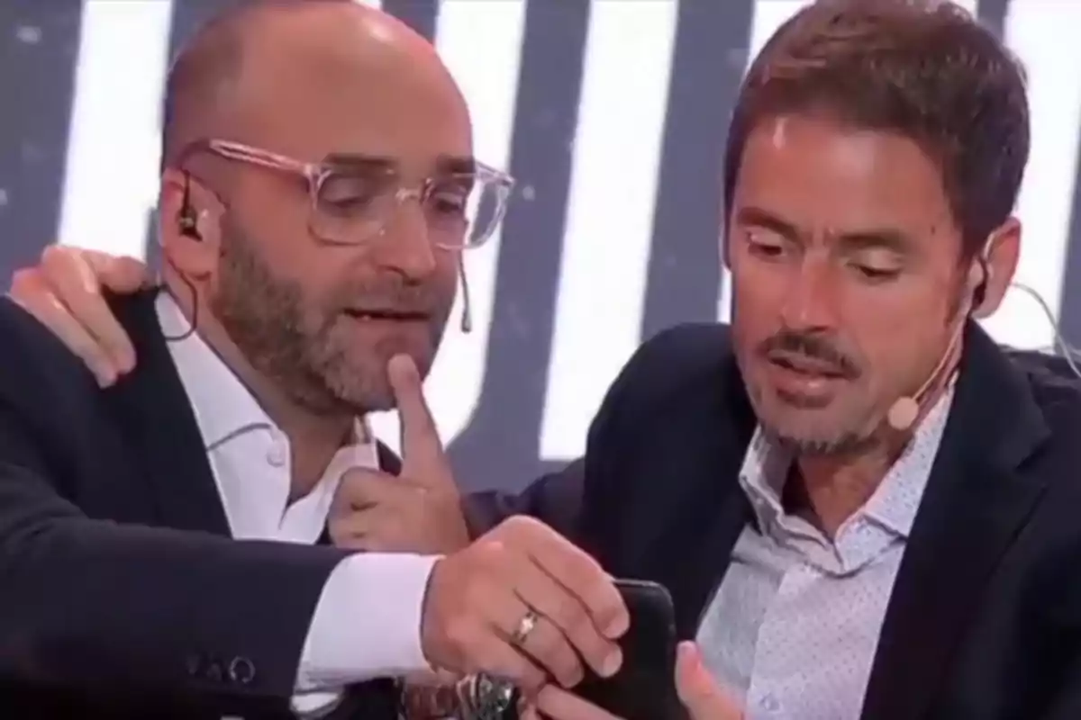 Two men with microphones and headphones closely watch a mobile phone while in an environment that appears to be a television studio.