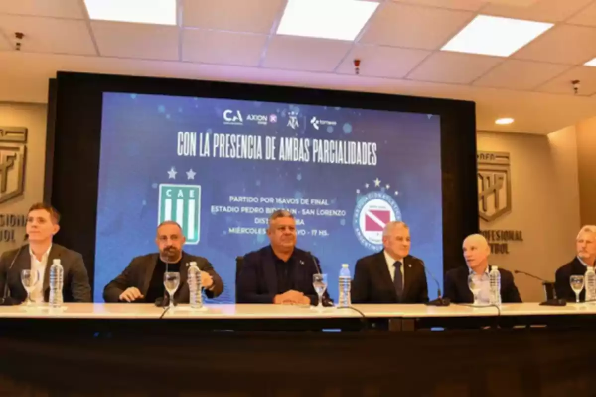 Seis hombres sentados en una mesa durante una conferencia de prensa con un cartel de fondo que anuncia un partido de fútbol entre dos equipos argentinos