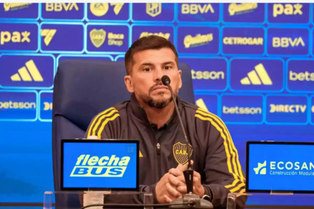 Un hombre en una conferencia de prensa con un micrófono frente a él y un fondo con logotipos de patrocinadores. Un hombre en una conferencia de prensa con un micrófono frente a él y un fondo con logotipos de patrocinadores.