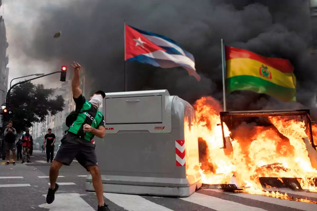 Una persona con el rostro cubierto lanza un objeto en una calle donde hay fuego y humo, con banderas de Cuba y Bolivia ondeando al fondo.