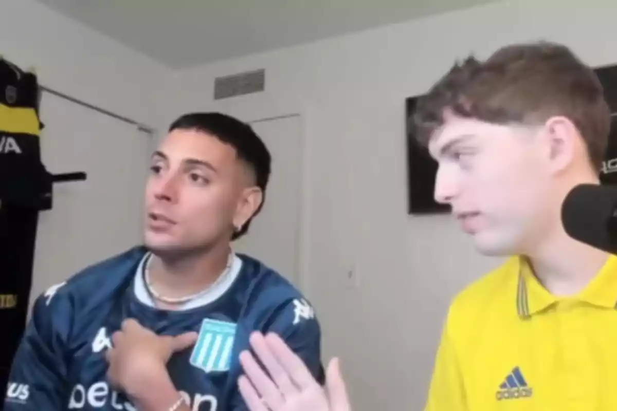 Two young men are talking in front of a microphone in a room; one is wearing a blue Racing jersey and the other a yellow Adidas jersey. In the background, a Boca Juniors jersey is hanging on the wall.