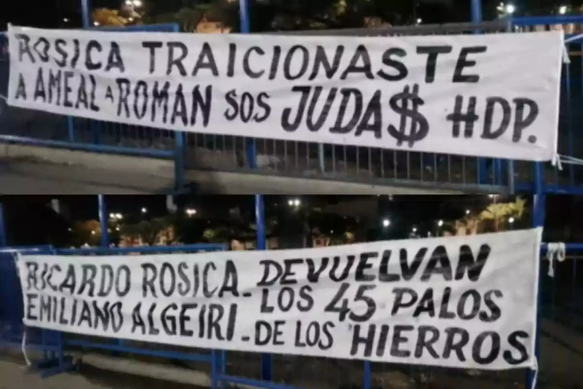 A banner with messages written in black letters on a white background, hung on a blue metal structure, with a nighttime setting and lights in the background. A banner with messages written in black letters on a white background, hung on a blue metal structure, with a nighttime setting and lights in the background.