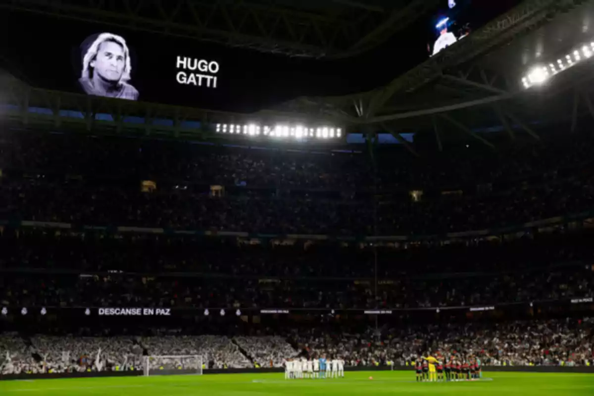 A soccer stadium full of spectators, with two teams on the field observing a minute of silence, while a black and white image of a person with the text A soccer stadium full of spectators, with two teams on the field observing a minute of silence, while a black and white image of a person with the text