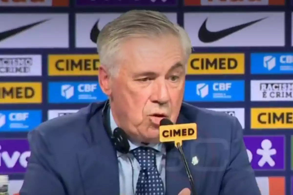 Gray-haired man in a blue suit speaking at a press conference with a microphone and a backdrop of sports sponsors Gray-haired man in a blue suit speaking at a press conference with a microphone and a backdrop of sports sponsors