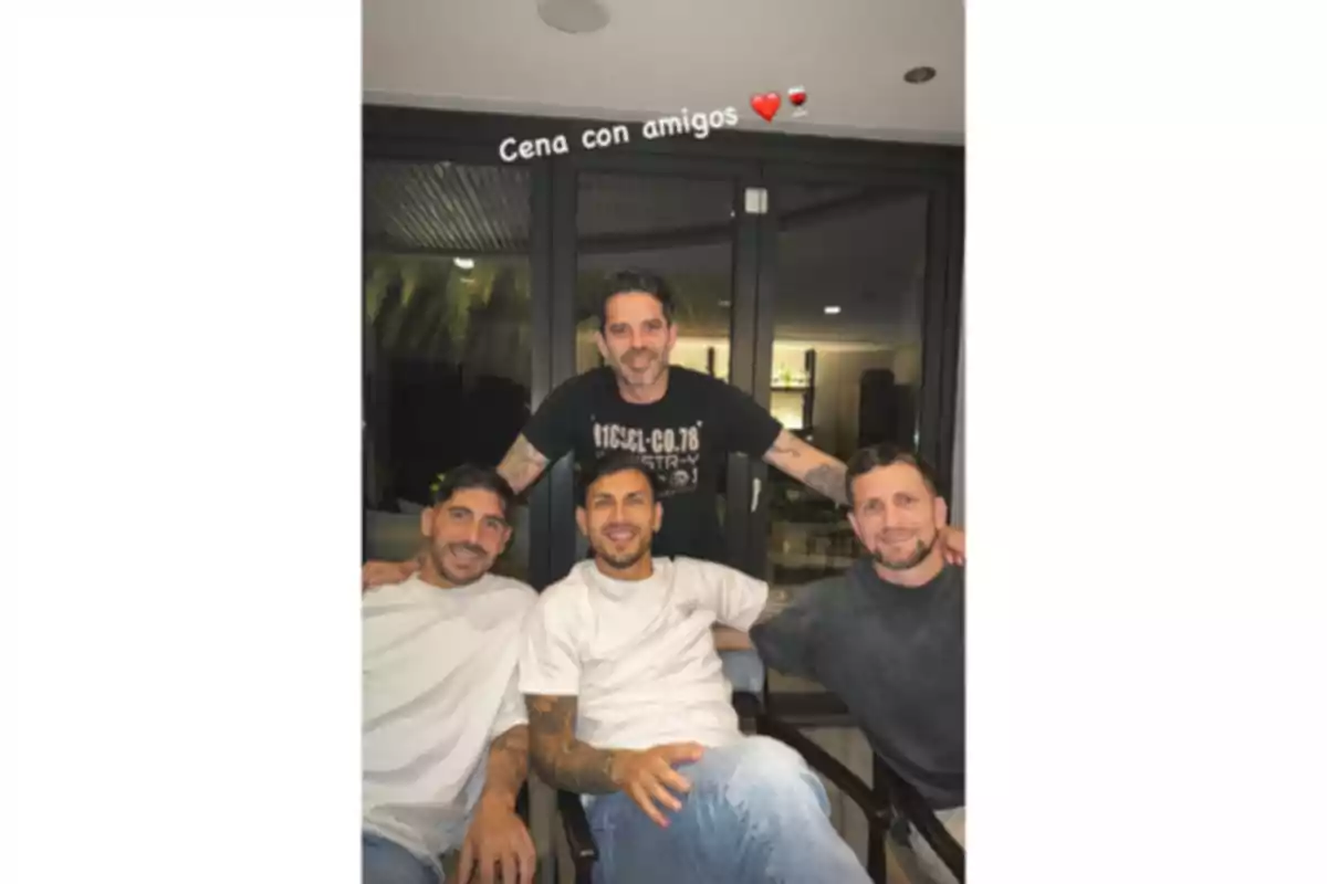 Cuatro amigos posan sonrientes en una cena, tres están sentados y uno de pie detrás de ellos, al fondo se ve una puerta de vidrio y la frase cena con amigos con emojis de corazón y copa de vino. Cuatro amigos posan sonrientes en una cena, tres están sentados y uno de pie detrás de ellos, al fondo se ve una puerta de vidrio y la frase cena con amigos con emojis de corazón y copa de vino.