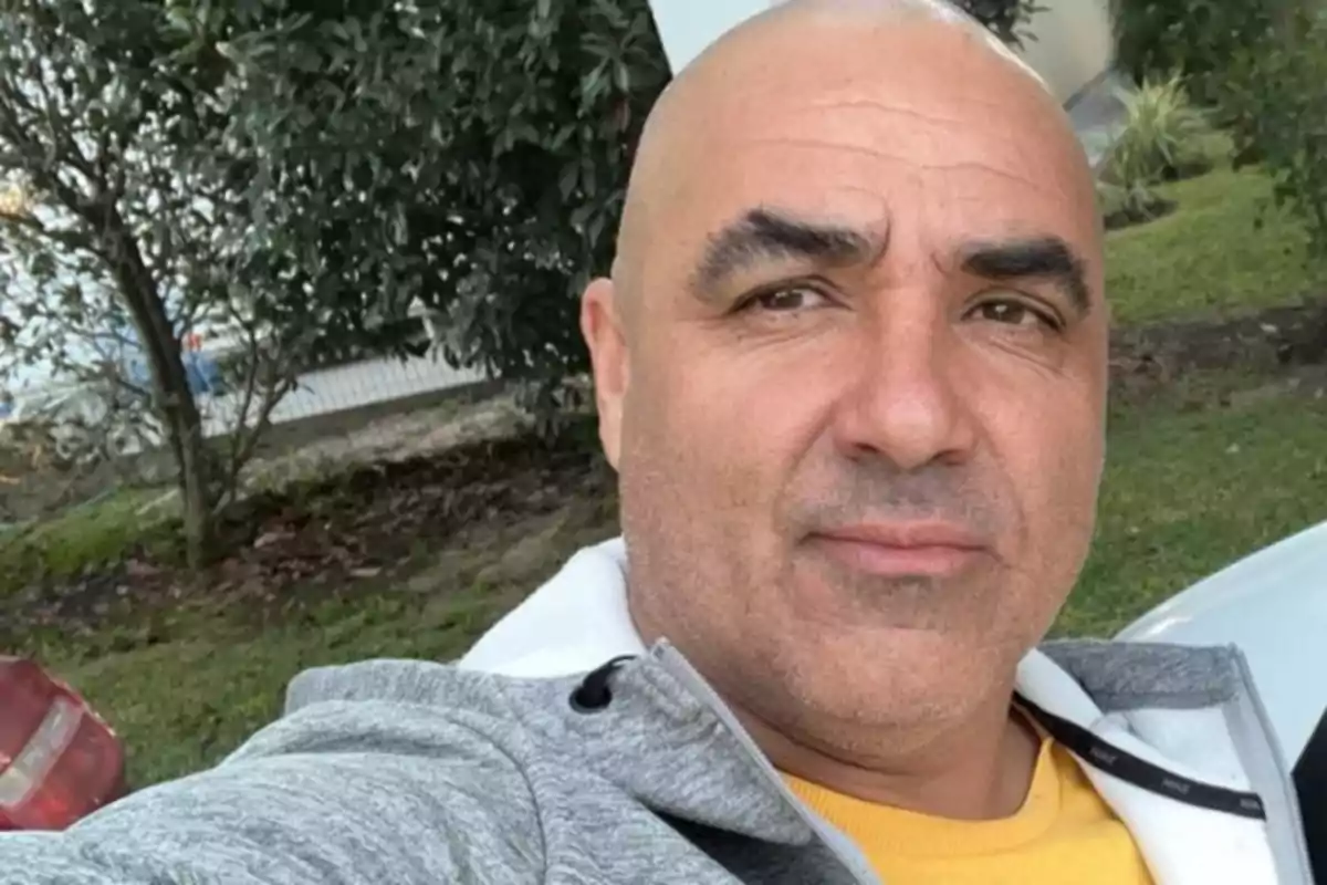 Bald man wearing a gray sweatjersey and a yellow t-jersey taking a selfie outdoors with trees and grass in the background