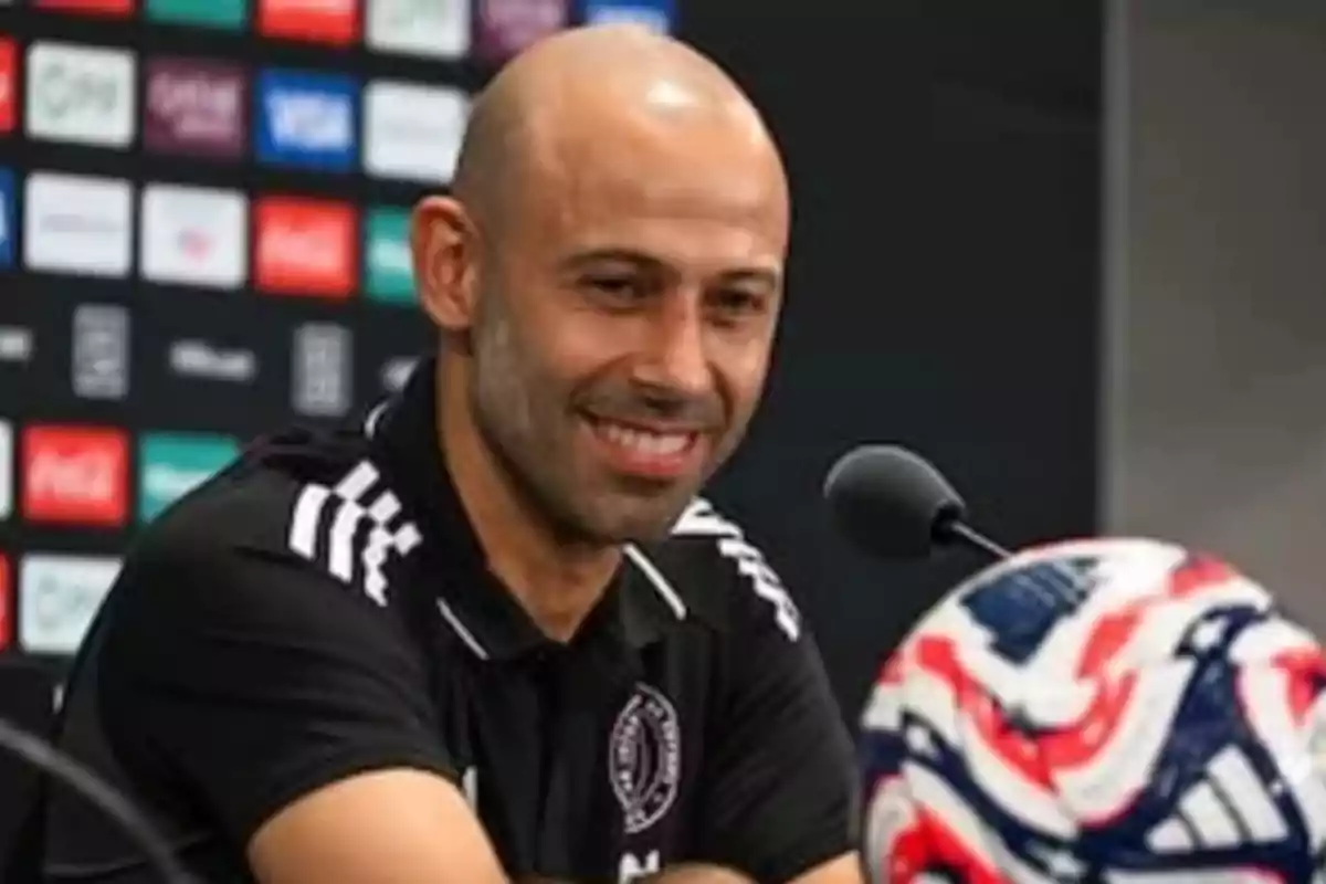 Bald man with a short beard smiling during a sports press conference with a microphone and a soccer ball in the foreground
