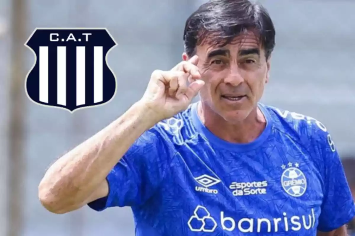 A man in a blue Grêmio jersey gestures with his hand next to the Club Atlético Talleres crest. A man in a blue Grêmio jersey gestures with his hand next to the Club Atlético Talleres crest.
