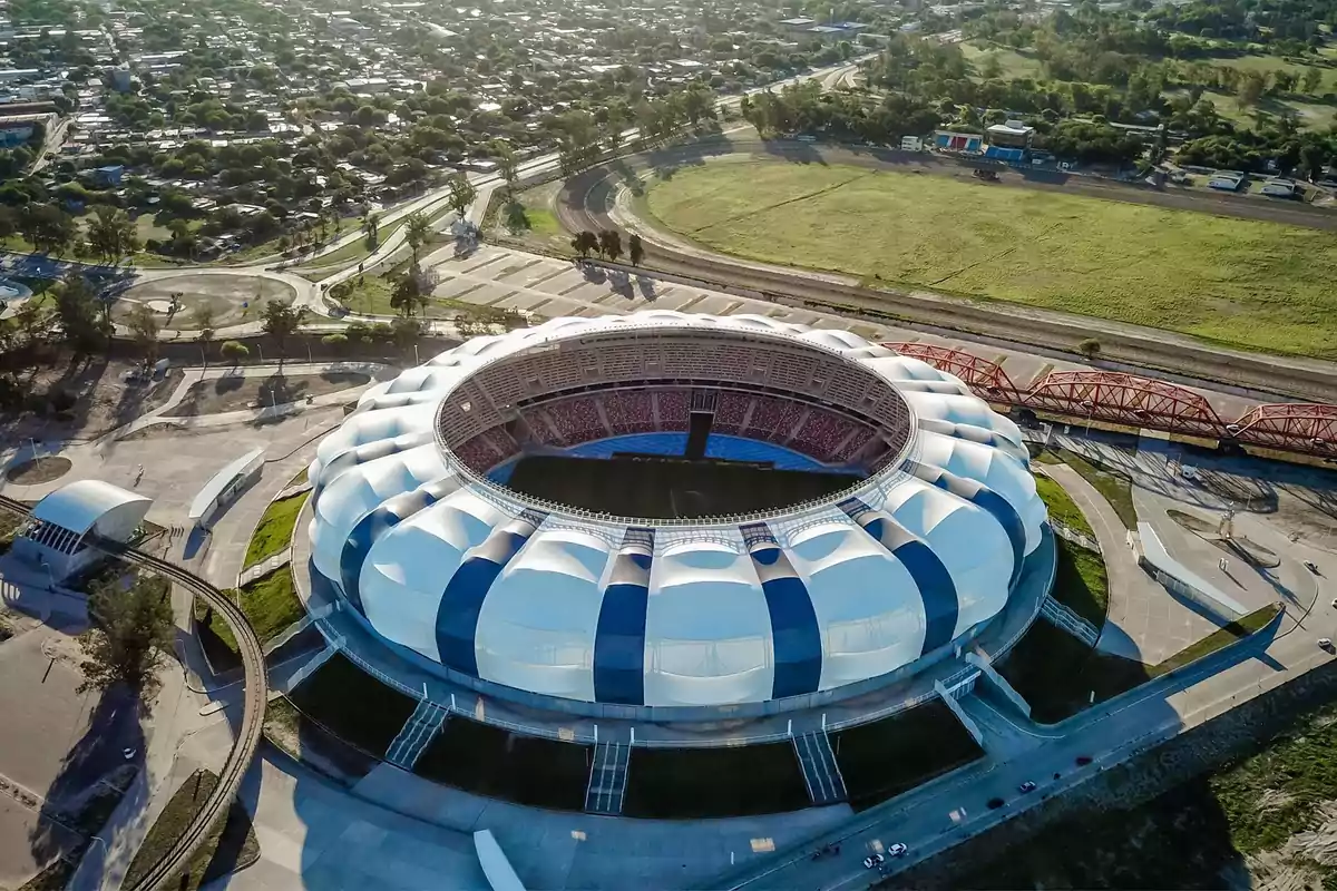 Aerial view of a modern stadium with a white and blue roof, surrounded by green areas and pathways in a city Aerial view of a modern stadium with a white and blue roof, surrounded by green areas and pathways in a city