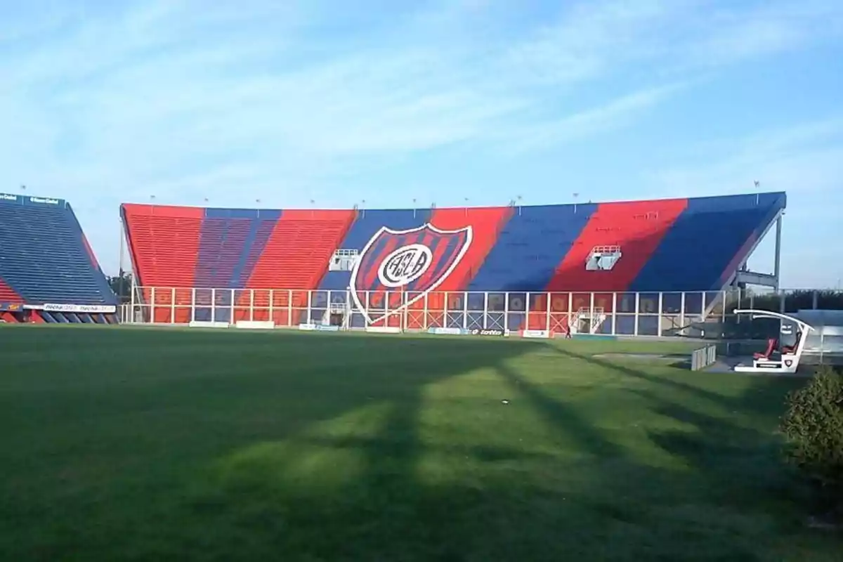 Un estadio de fútbol con gradas pintadas en rojo y azul y un escudo grande en el centro.