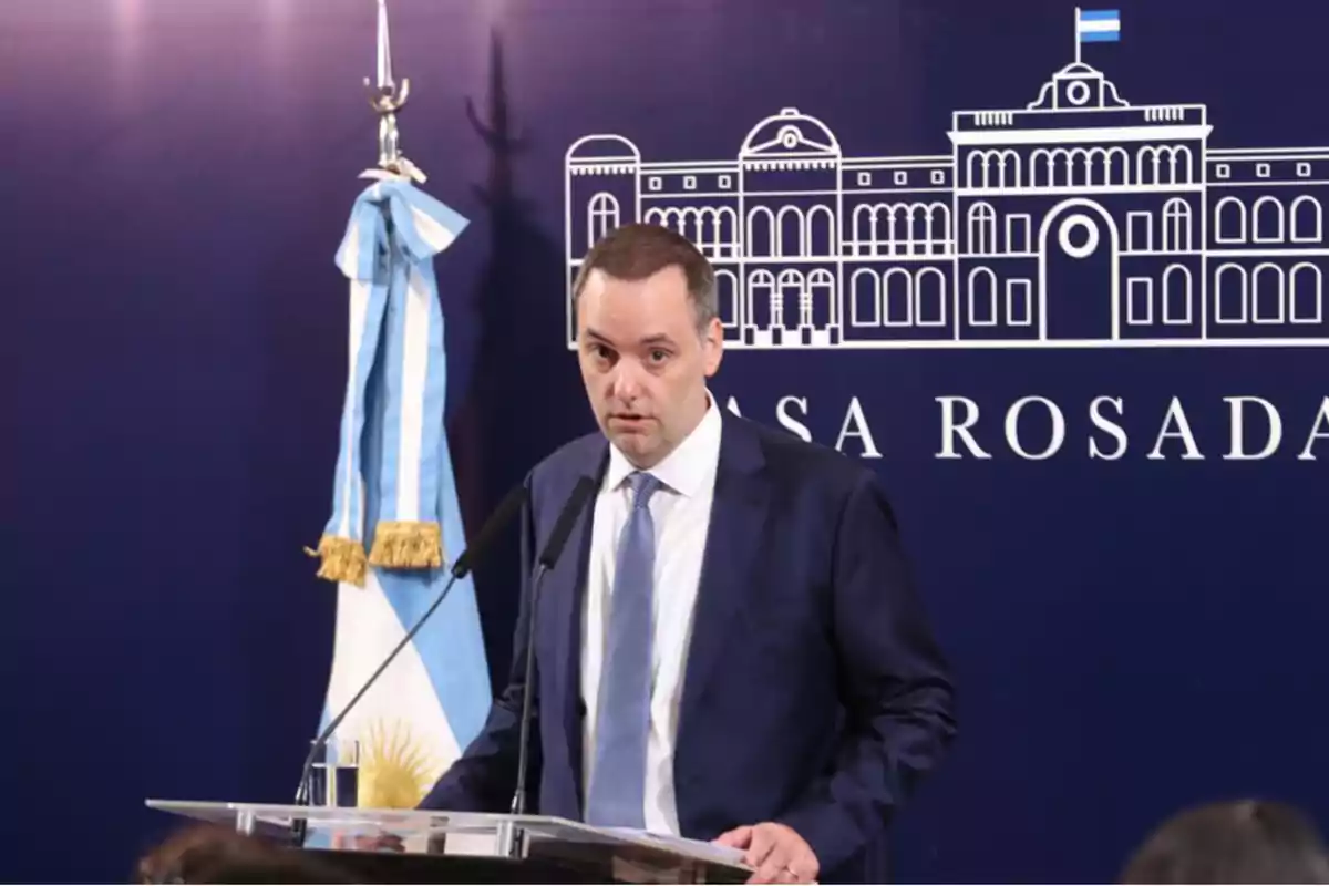 Un hombre en traje habla en un podio frente a un fondo azul con el dibujo de un edificio y una bandera argentina a su lado. Un hombre en traje habla en un podio frente a un fondo azul con el dibujo de un edificio y una bandera argentina a su lado.