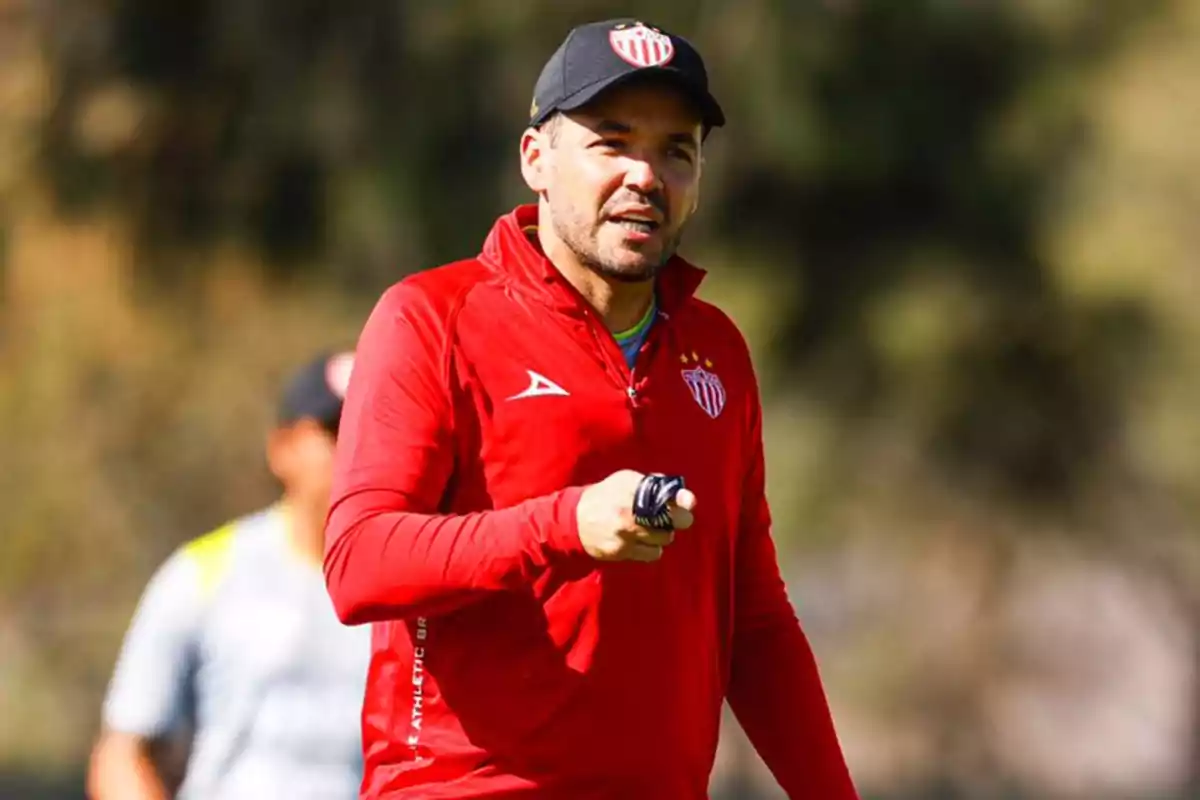 Entrenador de fútbol con chamarra y gorra rojas del Club Necaxa sosteniendo un silbato en un campo de entrenamiento Entrenador de fútbol con chamarra y gorra rojas del Club Necaxa sosteniendo un silbato en un campo de entrenamiento