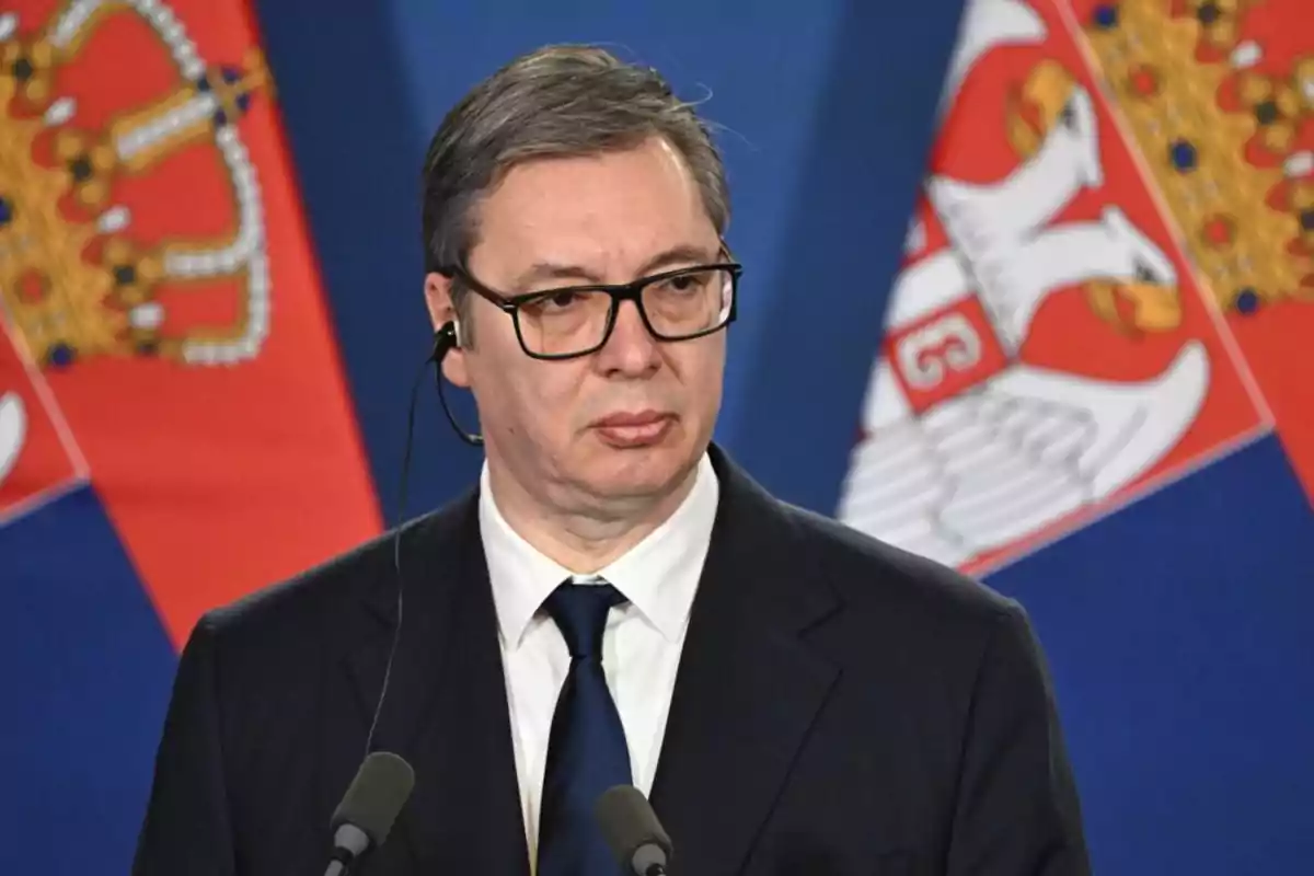 A man with glasses and headphones, dressed in a dark suit and tie, in front of microphones and flags with emblems. A man with glasses and headphones, dressed in a dark suit and tie, in front of microphones and flags with emblems.