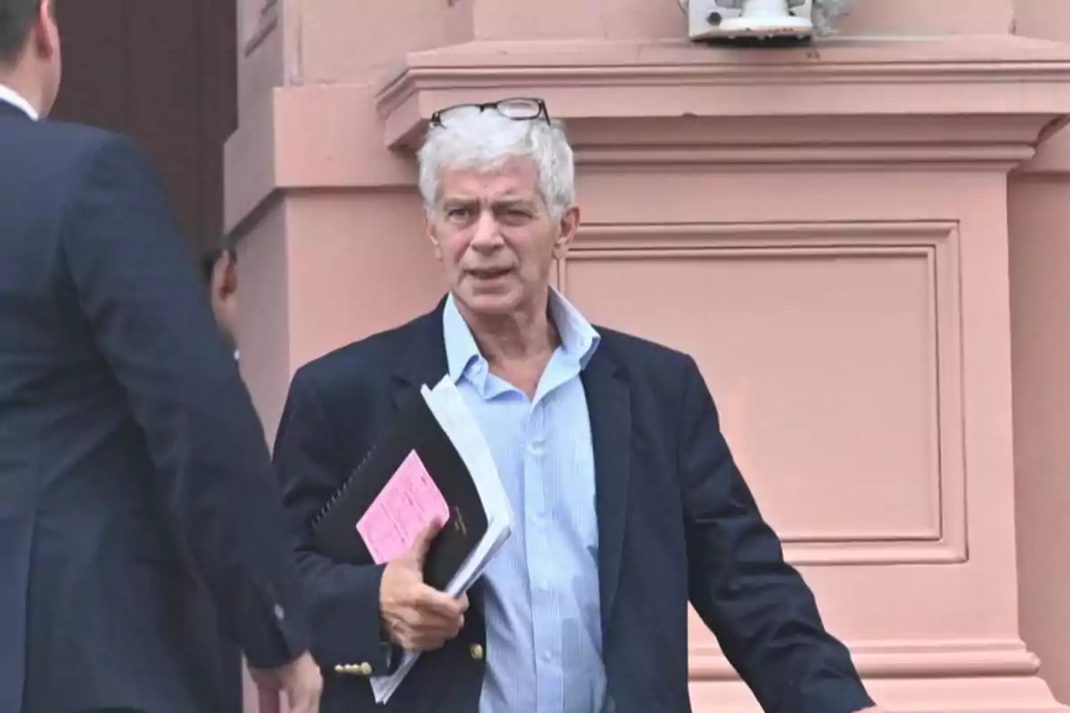 A gray-haired man with glasses on his head holds documents while walking next to another person in front of a pink building. A gray-haired man with glasses on his head holds documents while walking next to another person in front of a pink building.