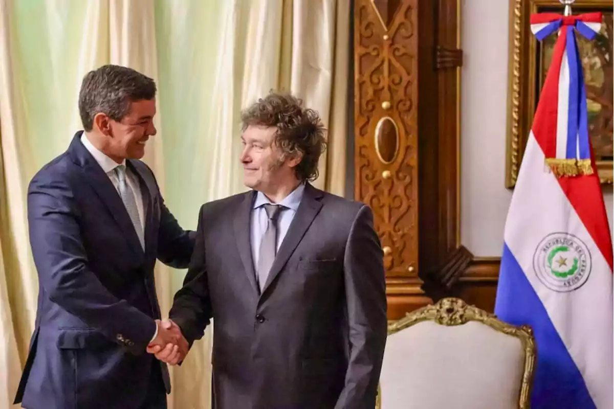 Dos hombres de traje se dan la mano en una sala decorada con una bandera de Paraguay.