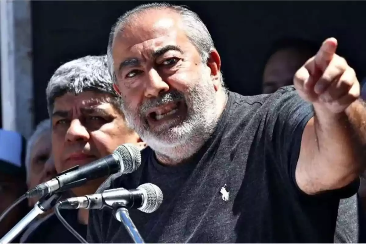Gray-bearded man speaking passionately in front of microphones and pointing with his finger while another person watches him from behind Gray-bearded man speaking passionately in front of microphones and pointing with his finger while another person watches him from behind