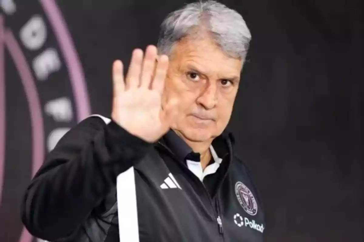 Un hombre de cabello canoso y chaqueta deportiva negra levanta la mano en señal de saludo.