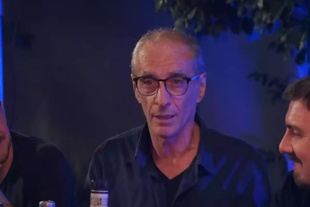 Un hombre con gafas y cabello canoso está sentado en una mesa junto a otras personas en un ambiente iluminado con luces azules.