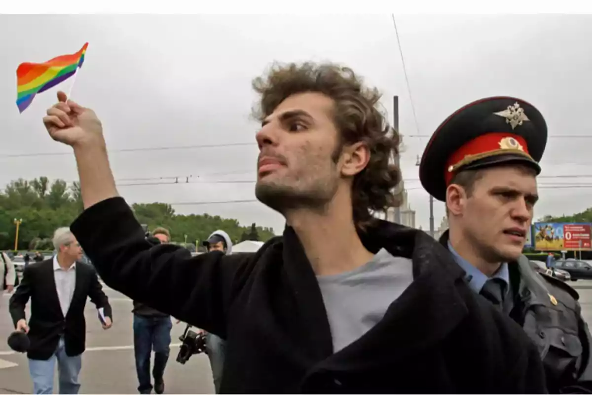 Un hombre sostiene una bandera arcoíris mientras es escoltado por un oficial de policía en un evento al aire libre.