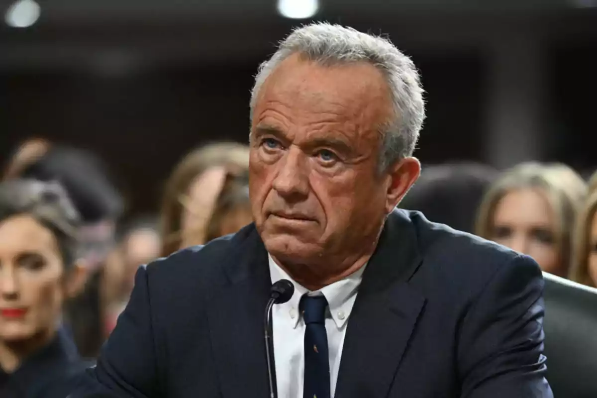 Hombre de cabello canoso con expresión seria usando traje oscuro y corbata, sentado frente a un micrófono en un entorno formal con personas al fondo desenfocadas