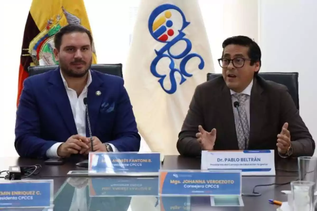 Two men in suits sitting at a conference table, with an Ecuadorian flag and an institutional flag with the CPCCS logo visible in the background.