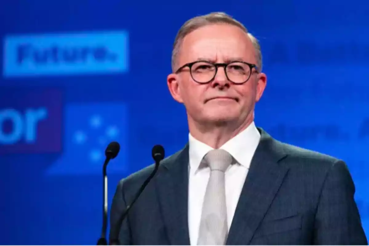 A man with glasses and a suit speaks in front of a microphone with a blue background. A man with glasses and a suit speaks in front of a microphone with a blue background.