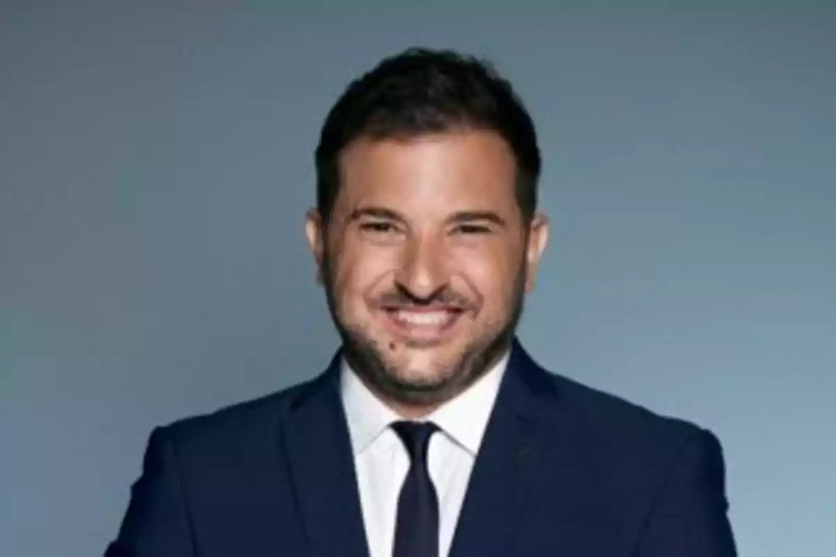 Hombre sonriendo con traje oscuro y corbata sobre fondo gris.