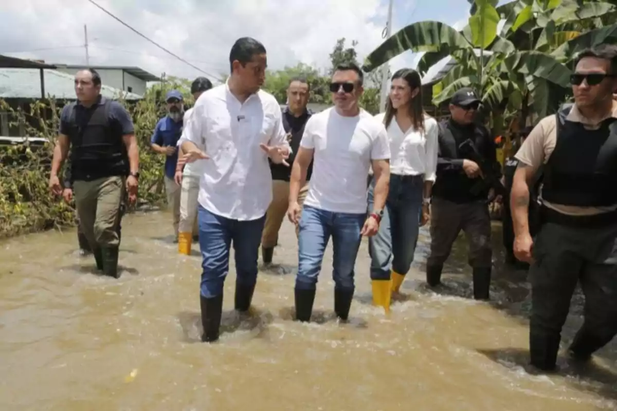 A group of people walks through a flooded area, some wearing rubber boots and surrounded by vegetation.