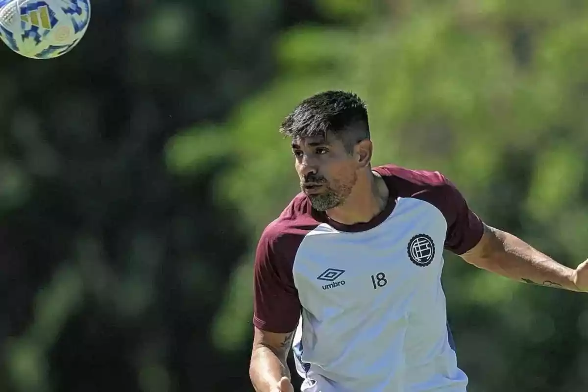 Un jugador de fútbol con camiseta blanca y detalles marrones observa un balón en el aire durante un entrenamiento al aire libre.