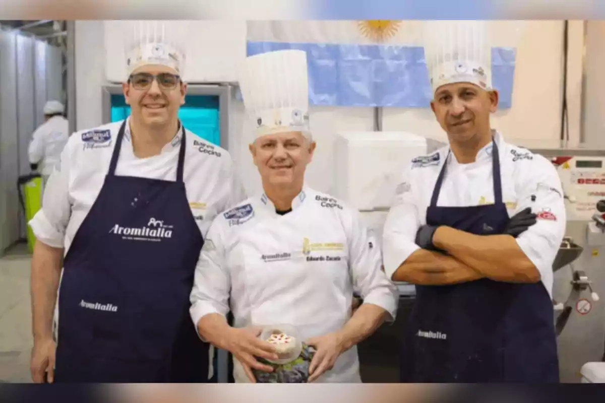 Tres chefs con gorros altos y delantales posan juntos en una cocina profesional con una bandera de Argentina de fondo Tres chefs con gorros altos y delantales posan juntos en una cocina profesional con una bandera de Argentina de fondo