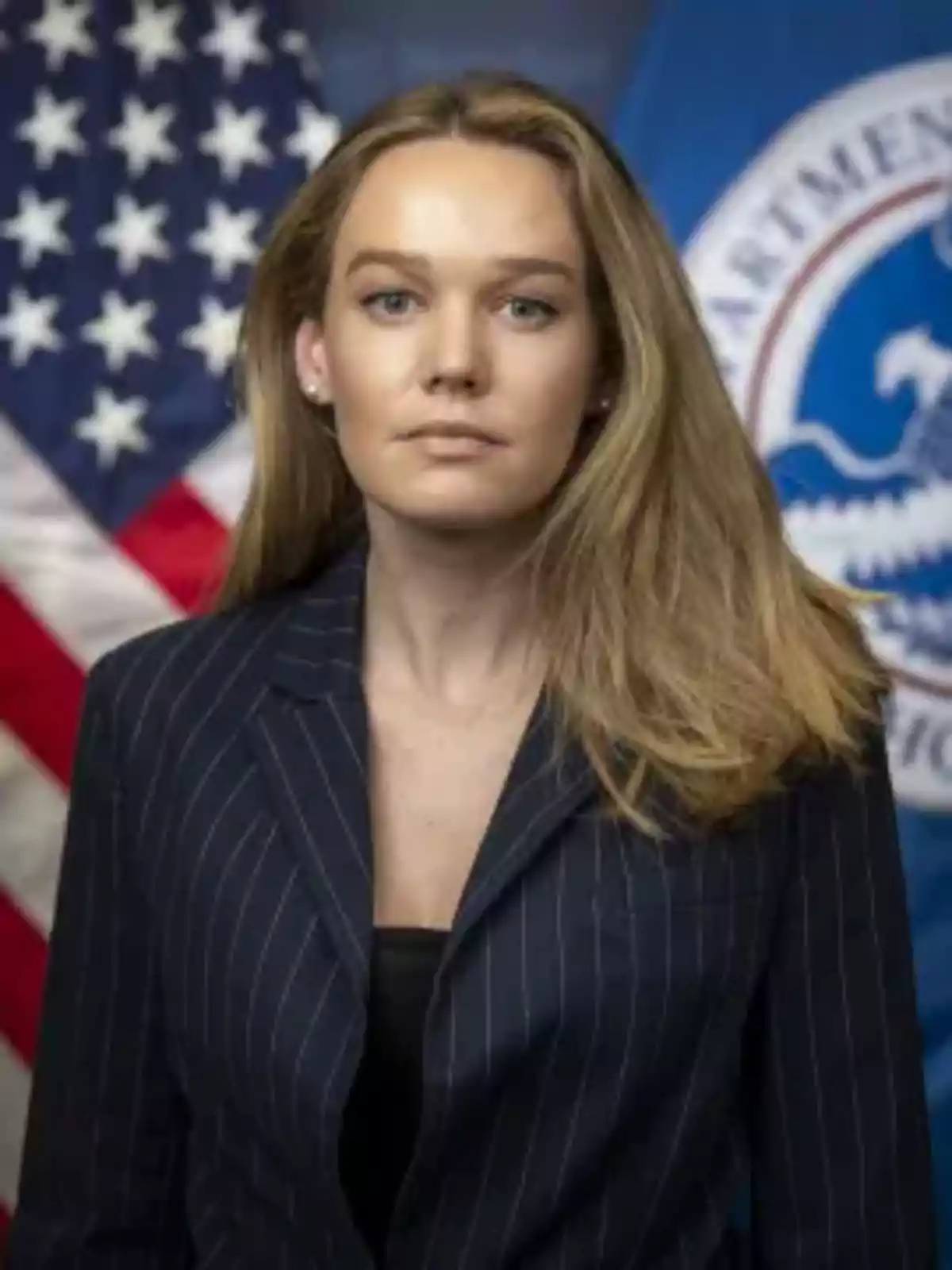 Blonde, straight-haired woman wearing a dark striped blazer posing in front of a United States flag and a blue institutional background Blonde, straight-haired woman wearing a dark striped blazer posing in front of a United States flag and a blue institutional background