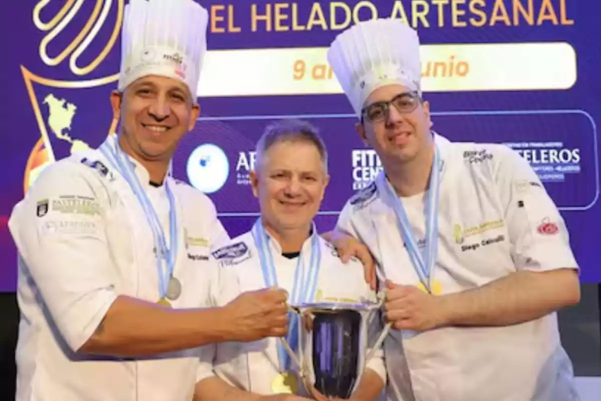 Tres chefs con uniformes y gorros blancos sonríen mientras sostienen un trofeo y llevan medallas en el cuello frente a un fondo con texto sobre helado artesanal.