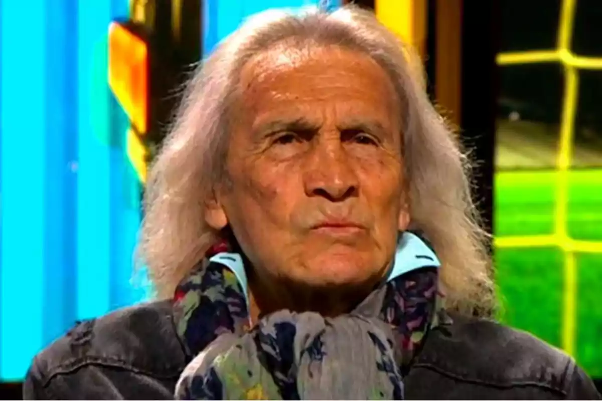 A man with long, gray hair, a colorful scarf, and a serious expression, set against a colorful and bright background. A man with long, gray hair, a colorful scarf, and a serious expression, set against a colorful and bright background.
