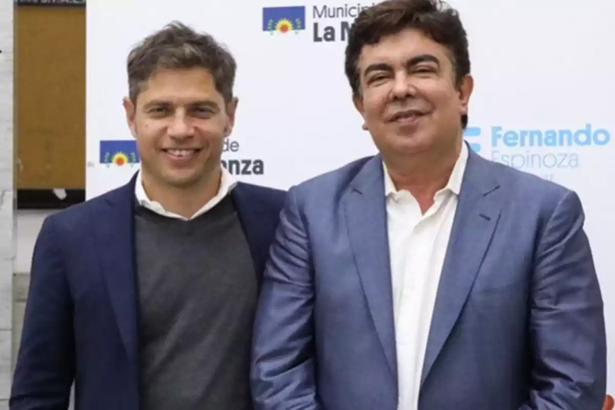 Dos hombres de traje posan juntos y sonríen frente a un fondo con logotipos institucionales.