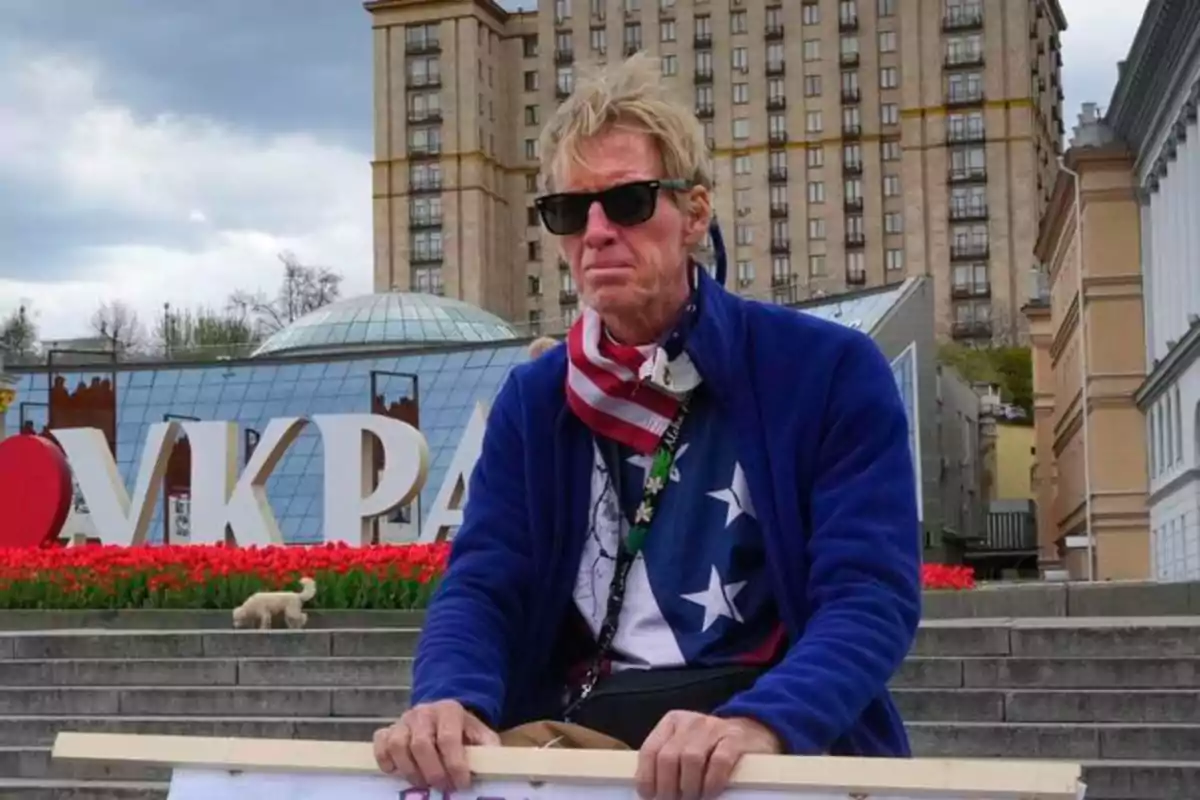 Un hombre con gafas de sol y bufanda está sentado frente a un edificio con un letrero grande y flores rojas.